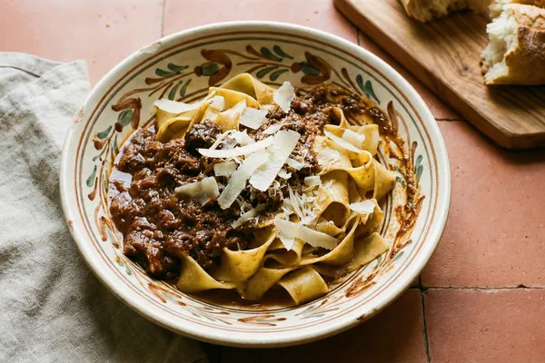 Wide pappardelle ribbons with dark wild boar ragu and grated cheese in a hand-painted ceramic bowl on terracotta