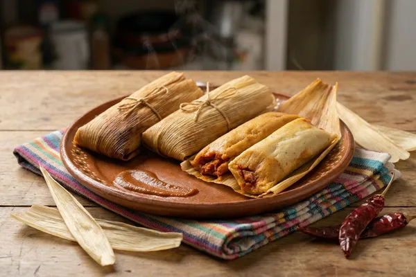 Steamed tamales on an earthenware plate, two unwrapped showing red chile pork filling inside golden masa